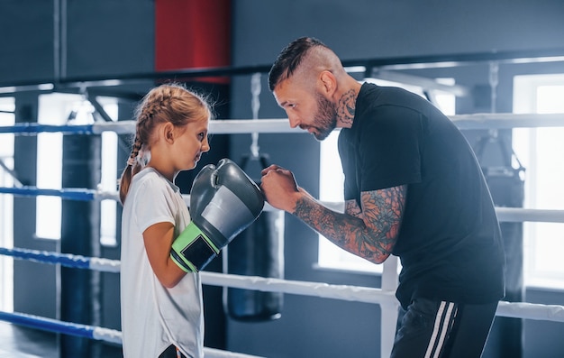 Avoir du sparring les uns avec les autres sur le ring de boxe. Le jeune entraîneur de boxe tatoué enseigne la petite fille mignonne dans le gymnase.
