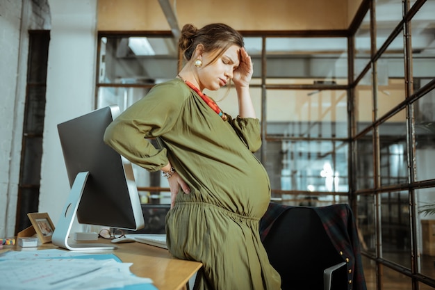 Avoir mal à la tête. Femme enceinte aux cheveux noirs vêtue d'une robe kaki ayant des maux de tête après avoir travaillé