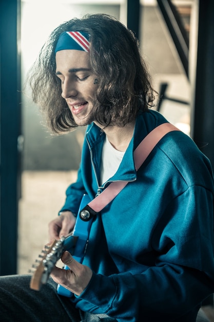 Avoir une répétition personnelle. Bel homme inspiré avec un tatouage sur le visage chantant les yeux fermés tout en accompagnant de la guitare