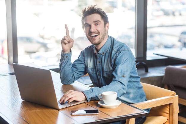 Ayez une idée ! Portrait d'un jeune pigiste barbu créatif et positif en chemise de jean bleu assis dans un café et travaillant sur un ordinateur portable avec un sourire à pleines dents et montrant le doigt vers le haut, regardant la caméra. intérieur