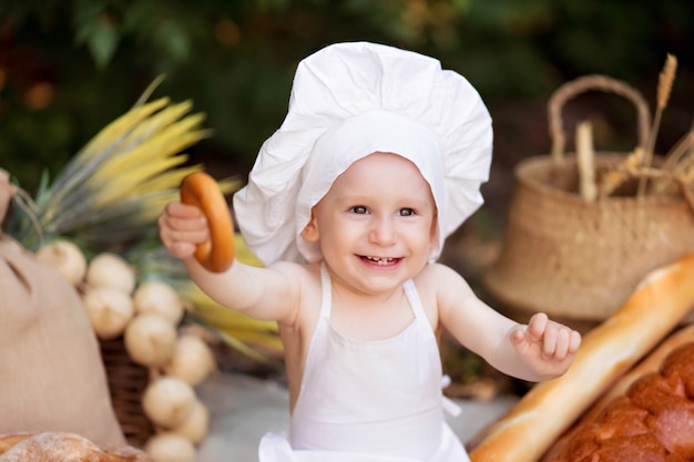 Baby baker cuisine dans la nature. Garçon sur un pique-nique mange du pain et des bagels dans un tablier blanc et un chapeau