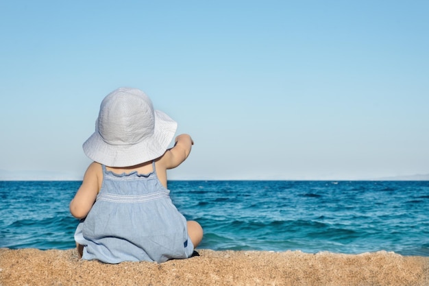 Baby Girl wearing sunhat,assis sur une plage de sable,regardez la mer,tourné de dos.Copy space