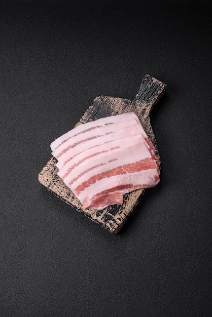 Bacon cru frais coupé en tranches avec des épices et des herbes salées sur un fond de béton texturé foncé