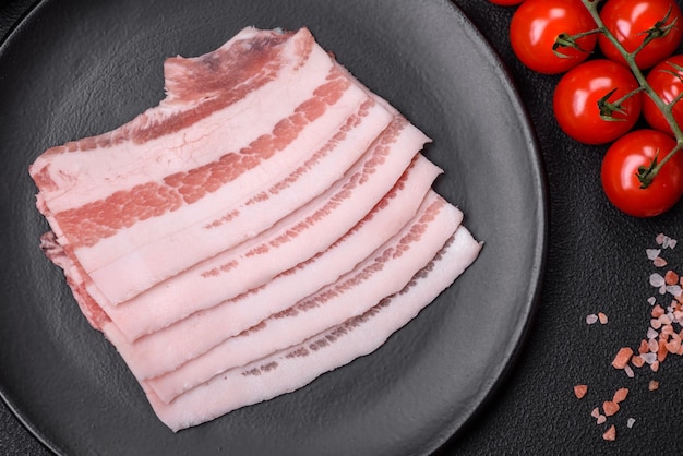 Bacon cru frais coupé en tranches avec des épices et des herbes salées sur un fond de béton texturé foncé