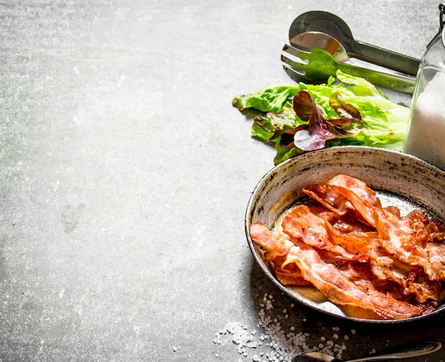 Bacon dans une poêle avec du sel et des herbes. Sur un fond de pierre.