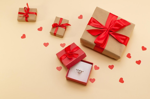 Une bague avec une grosse pierre dans une boîte à côté de cadeaux avec des rubans rouges sur fond beige avec des coeurs