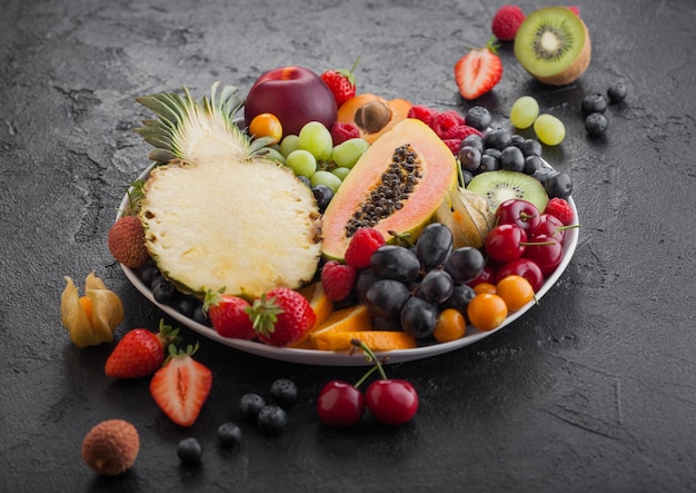 Baies d'été biologiques crues fraîches et fruits exotiques en plaque blanche sur fond noir Ananas papaye raisins nectarine orange abricot kiwi poire litchis cerise et physalis Vue de dessus