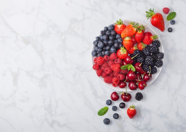 Photo les baies d'été biologiques fraîches se mélangent dans une assiette blanche sur fond de marbre. framboises, fraises, myrtilles, mûres et cerises. vue de dessus