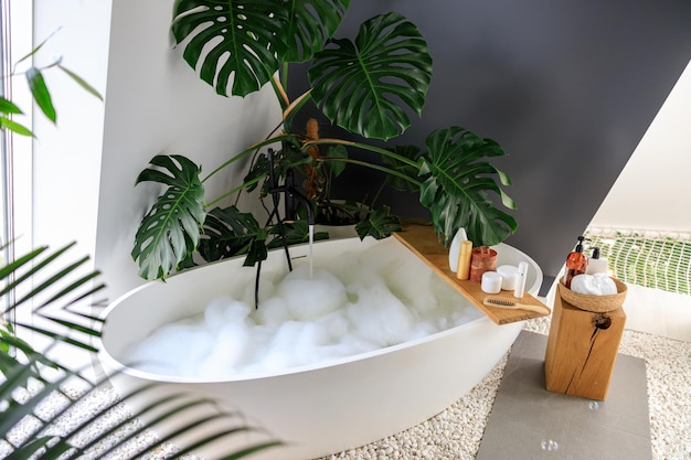 Baignoire remplie de bulles dans la salle de bain décorée de produits de soins corporels écologiques à base de plantes tropicales monstera