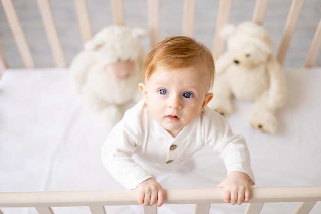 Le bâillement bébé garçon blond de 6 mois avec un jouet dans ses mains sur un lit blanc dans une chambre lumineuse dans un body en coton veut dormir