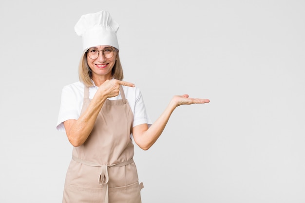 Baker d'âge moyen femme souriant joyeusement et pointant pour copier l'espace sur la paume sur le côté, montrant ou annonçant un objet contre un mur plat