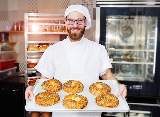 Baker attrayant en uniforme blanc tenant un plateau avec des bagels fraîchement cuits au four avec des graines de sésame et de pavot à la surface d'une boulangerie ou d'une boulangerie