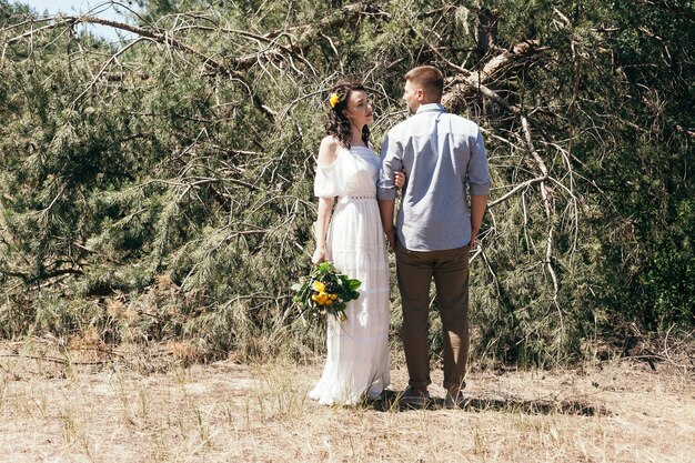 Balade nuptiale dans la pinède. Journée ensoleillée. Couple de mariage dans la forêt. Belle mariée et le marié en promenade. Robe de mariée blanche. Bouquet de pivoines et d'hortensias.