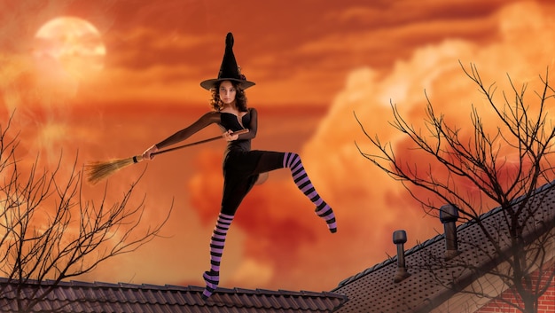 Une ballerine en chaussures de pointe dans un costume de sorcière noire avec un chapeau et un balai danse sur le toit d'une vieille maison sur fond d'un coucher de soleil lumineux et sinistre