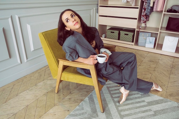 Ballerine en costume rayé gris avec une tasse de café souriant et rêvant est assise dans un fauteuil