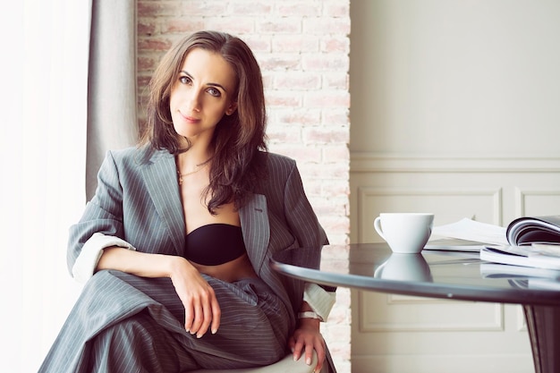 Ballerine en costume rayé gris avec une tasse de café souriant et rêvant est assise à une table près de la fenêtre