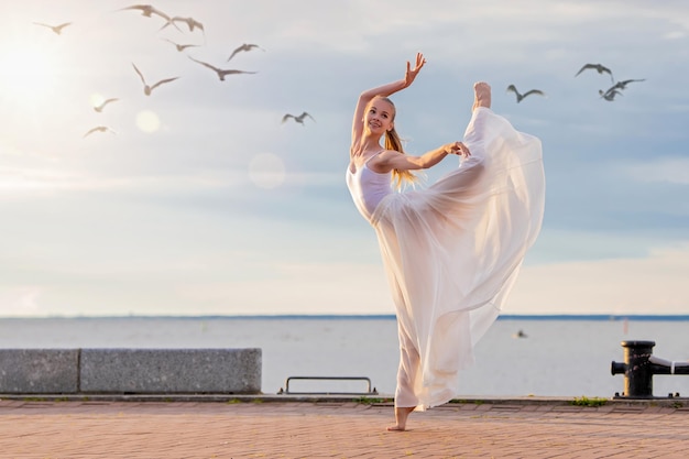 Une ballerine dansante dans une jupe volante blanche et des collants sur la digue de l'océan ou sur la plage de la mer entourée de mouettes dans le ciel