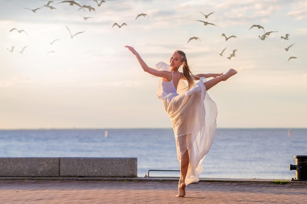 Une ballerine dansante dans une jupe volante blanche et des collants sur la digue de l'océan ou sur la plage de la mer entourée de mouettes dans le ciel