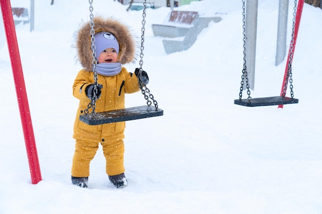 Photo un bambin heureux de 1217 mois en vêtements d'hiver jaunes tient sur une balançoire et aime tomber des flocons de neige tout en se tenant sur une aire de jeux pour enfants en hiver