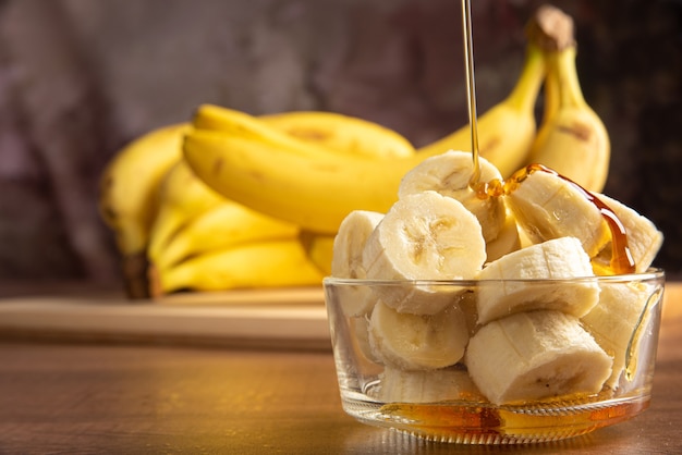 Banane coupée en tranches dans un bocal en verre baigné de miel avec plus de bananes en arrière-plan, fond abstrait sombre, mise au point sélective.