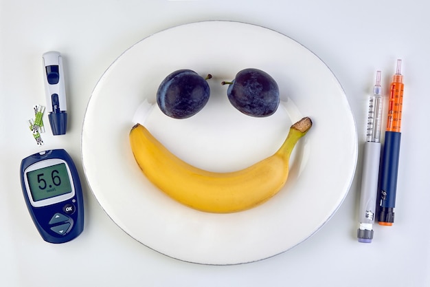 Banane et prunes sous la forme d'une émoticône souriante sur une plaque blanche, des stylos à seringue à insuline et un glucomètre comme couverts sur une table blanche Glycémie normale