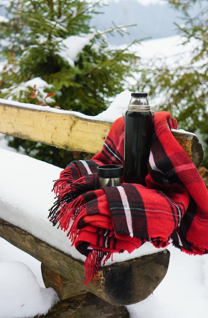 Banc en bois rustique rugueux avec plaid tartan rouge de Noël confortable et tasse thermos Arrière-cour rurale avec pin hiver dans la campagne par temps de neige en plein air