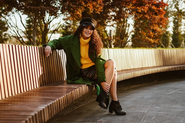 Sur un banc de parc avec son pied sur sa jambe se trouve une beauté aux cheveux bouclés vêtue d'un style militaire d'automne