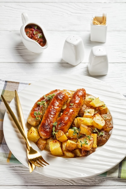 Bangers et pommes de terre rôties sur une assiette blanche avec sauce à l'oignon dans une cruche sur une table en bois texturée blanche, vue verticale d'en haut