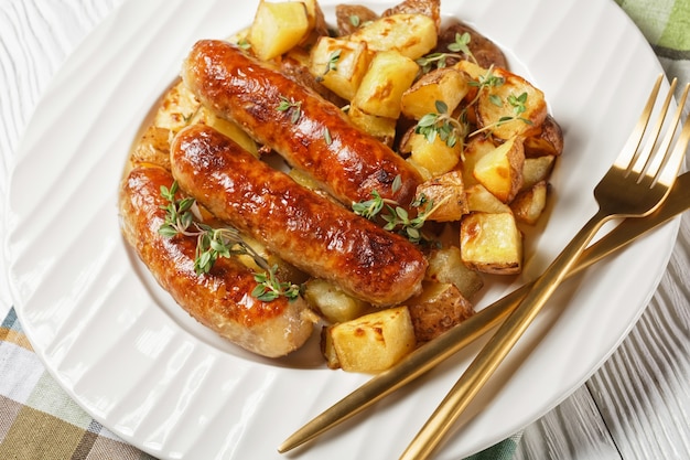 Bangers et pommes de terre rôties sur une assiette blanche avec sauce à l'oignon dans une cruche sur une table en bois texturée blanche