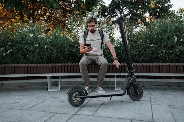 Banlieusard homme hipster avec scooter électrique à l'extérieur en ville à l'aide d'un smartphone