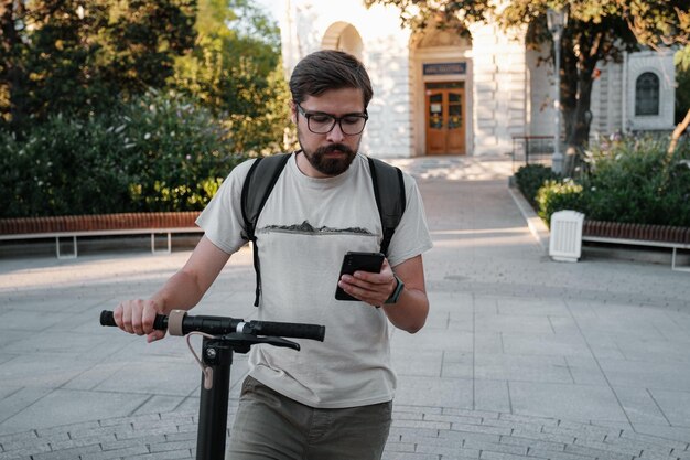 Banlieusard homme hipster avec scooter électrique à l'extérieur en ville à l'aide d'un smartphone