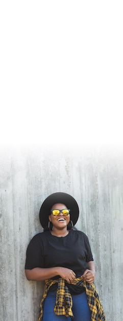 Bannière belle fille afro-américaine hipster millénaire regarde la caméra et souriant porte des lunettes de soleil et un chapeau tout en se tenant contre le mur espace de copie vertical et espace vide pour le texte
