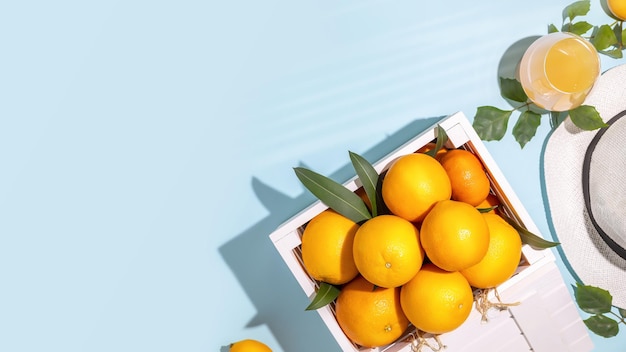 Bannière d'été avec des oranges de ferme avec des feuilles dans une boîte blanche en bois un verre de jus d'orange frais et un chapeau d'été sur un fond bleu avec espace de copie Fruits locaux de saison avec beaucoup de vitamines