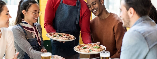 Bannière horizontale ou en-tête avec un serveur servant de délicieuses pizzas margherita à des amis multiculturels dans un restaurant pizzeria confortable Des amis multiethniques s'amusant ensemble à la pizzeria