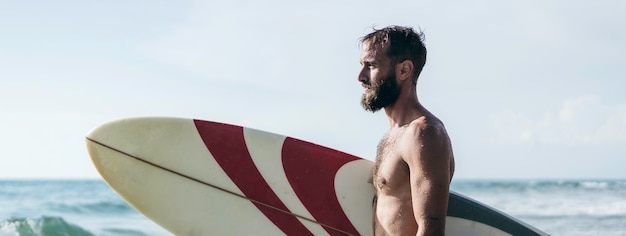 Bannière horizontale ou en-tête avec surfeur tenant sa planche de surf sur la plage Homme hipster debout sur la plage et attendant de grosses vagues pour surfer Homme barbu en forme avec planche de surf à la mer