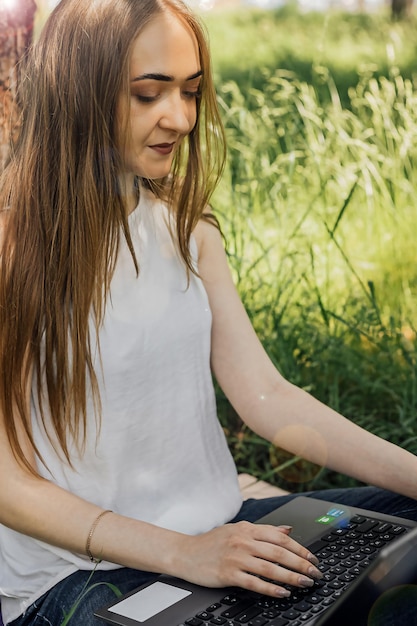 Sur la bannière, une jeune fille travaille avec un ordinateur portable à l'air frais dans le parc assis sur la pelouse Le concept de travail à distance Travailler en tant que pigiste La fille suit des cours sur un ordinateur portable et sourit