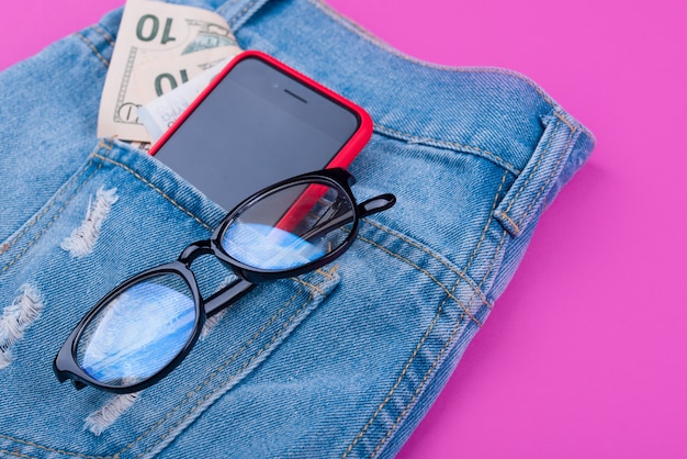 Bannière sur une surface rose avec un jean bleu, de l'argent, des écouteurs, un téléphone, des lunettes.