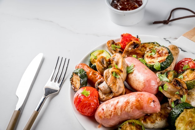 Barbecue. Assortiment de diverses saucisses de viande grillées, avec légumes BBQ - champignons, tomates, courgettes, oignons. Sur table en marbre blanc, sur assiette, avec sauce.