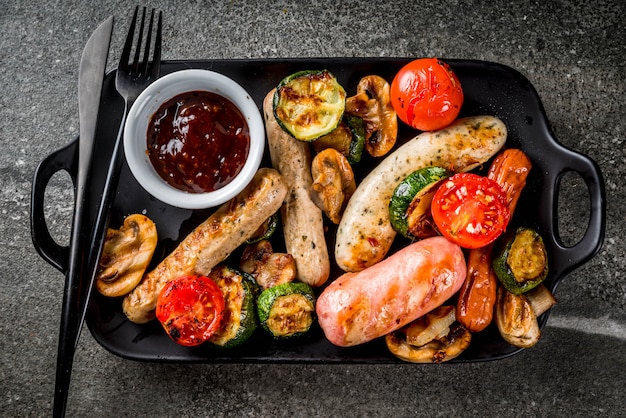 Barbecue. Assortiment de diverses saucisses de viande grillées, avec légumes BBQ champignons, tomates, courgettes, oignons. Sur une table en pierre noire, sur une assiette noire, avec sauce. vue de dessus du fond