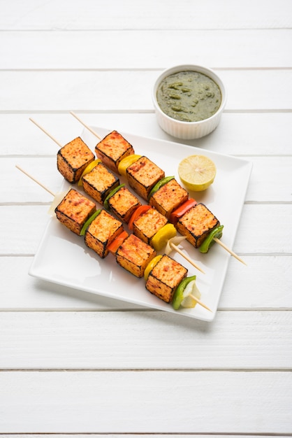 Barbecue Paneer Tikka Kabab - cubes de fromage cottage indien Tandoori en sauce rouge et blanche en bâtonnets, servis dans une assiette avec chutney vert sur une table colorée ou en bois, mise au point sélective