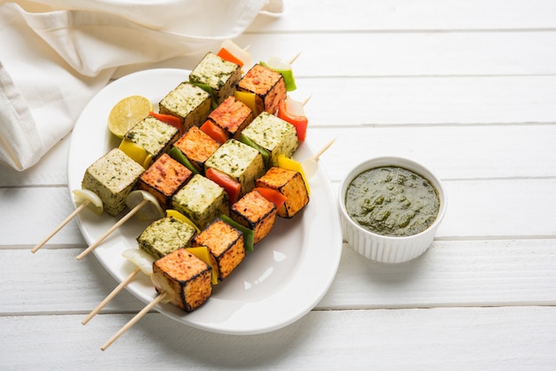 Barbecue Paneer Tikka Kabab - cubes de fromage cottage indien Tandoori en sauce rouge et blanche en bâtonnets, servis dans une assiette avec chutney vert sur une table colorée ou en bois, mise au point sélective