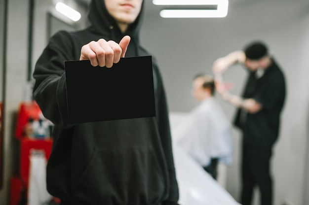 Barber in dark hoodie est titulaire d'une carte pour copyspace dans un salon de coiffure masculin et des clients de coupure de coiffeur