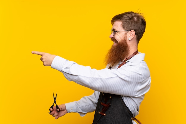 Photo barber avec une longue barbe dans un tablier sur un mur jaune isolé pointant sur le côté pour présenter un produit