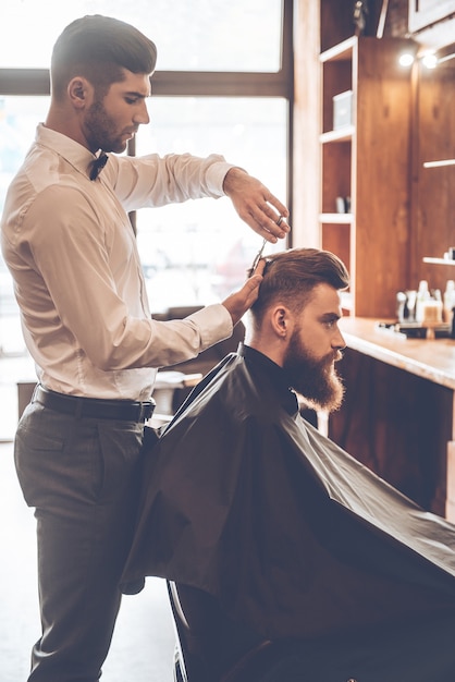 Barbier au travail. Vue latérale d'un jeune homme barbu se faisant couper les cheveux par un coiffeur assis sur une chaise au salon de coiffure