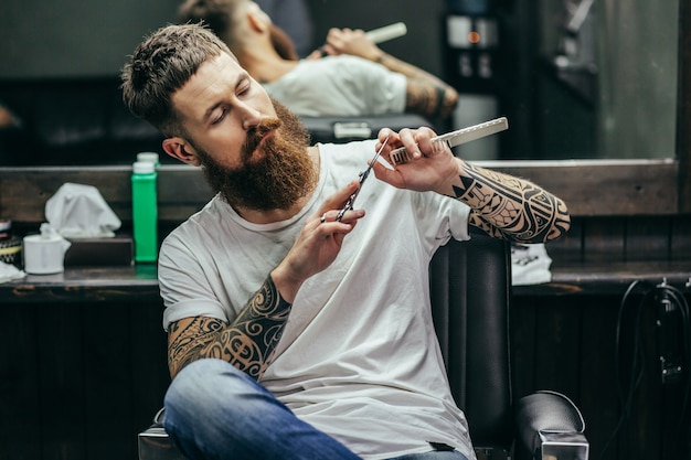 Barbier aux bras tatoués en regardant les ciseaux dans ses mains