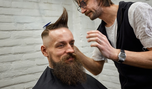 Barbier mâle fait une coiffure mohawk à l'aide de la tondeuse d'un homme adulte avec barbe