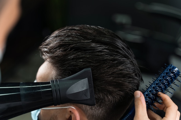 Barbier en masque médical sèche les cheveux d'un bel homme barbu après une coupe de cheveux à la mode. Le travail d'un coiffeur pendant la période de quarantaine coronavirus covid-19.