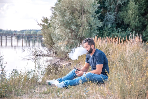 Barbu élégant fumant une vape sur le sol de la forêt. La cigarette nouvelle technologie.