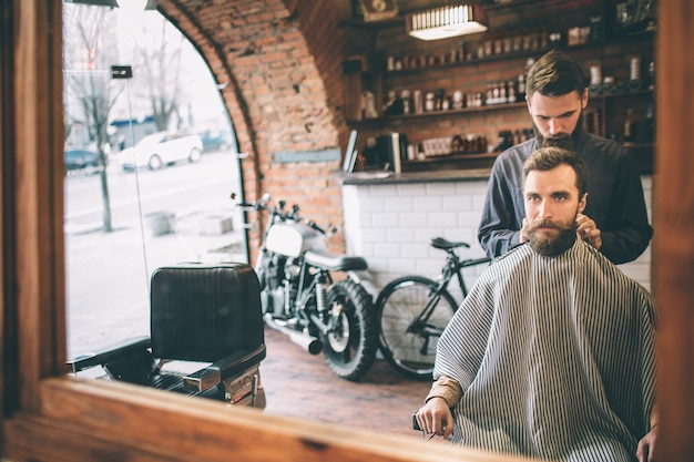 Un barbu est assis sur une chaise et regarde le miroir. Son coiffeur le prépare à couper des cheveux.