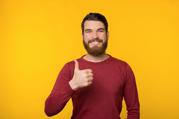 Barbu gai jeune homme en pull rouge montre le pouce vers le haut un sourire près d'un mur jaune.